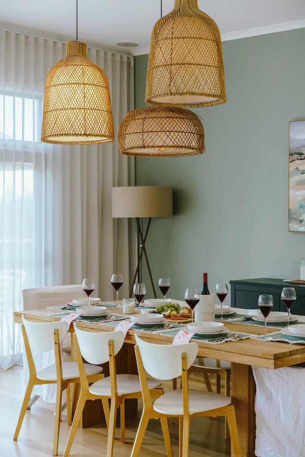 Dining table set with plates, wine glasses, and a bottle under three woven pendant lights.