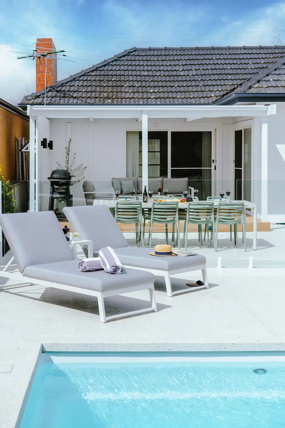Two gray lounge chairs by a pool with towels, hat, and table set outside a white house patio.