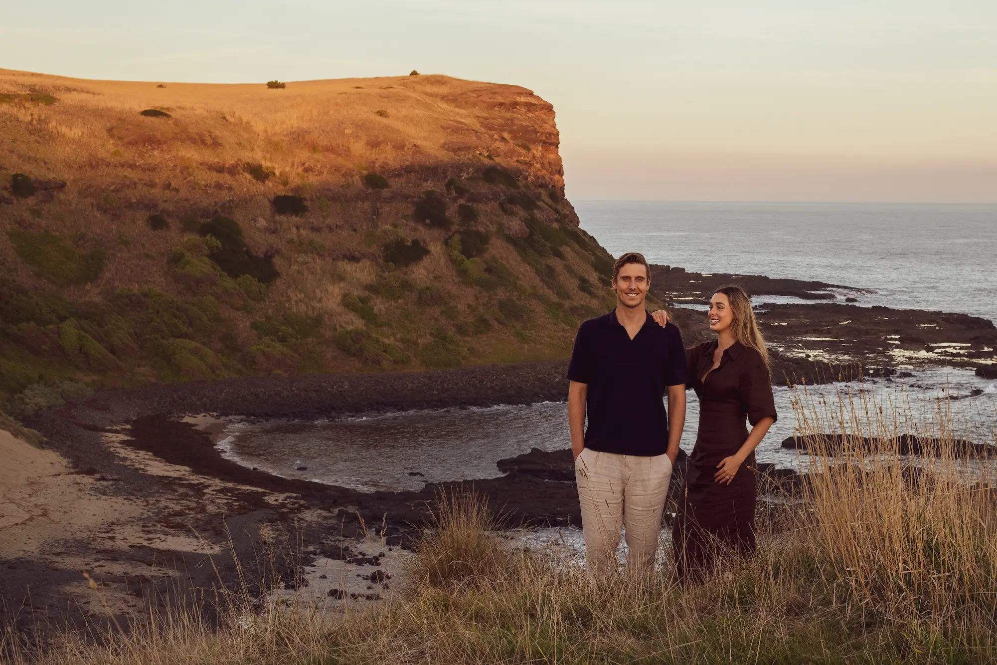 Man and woman standing on grassy cliffside with ocean and rocky coastline in the background at sunset.