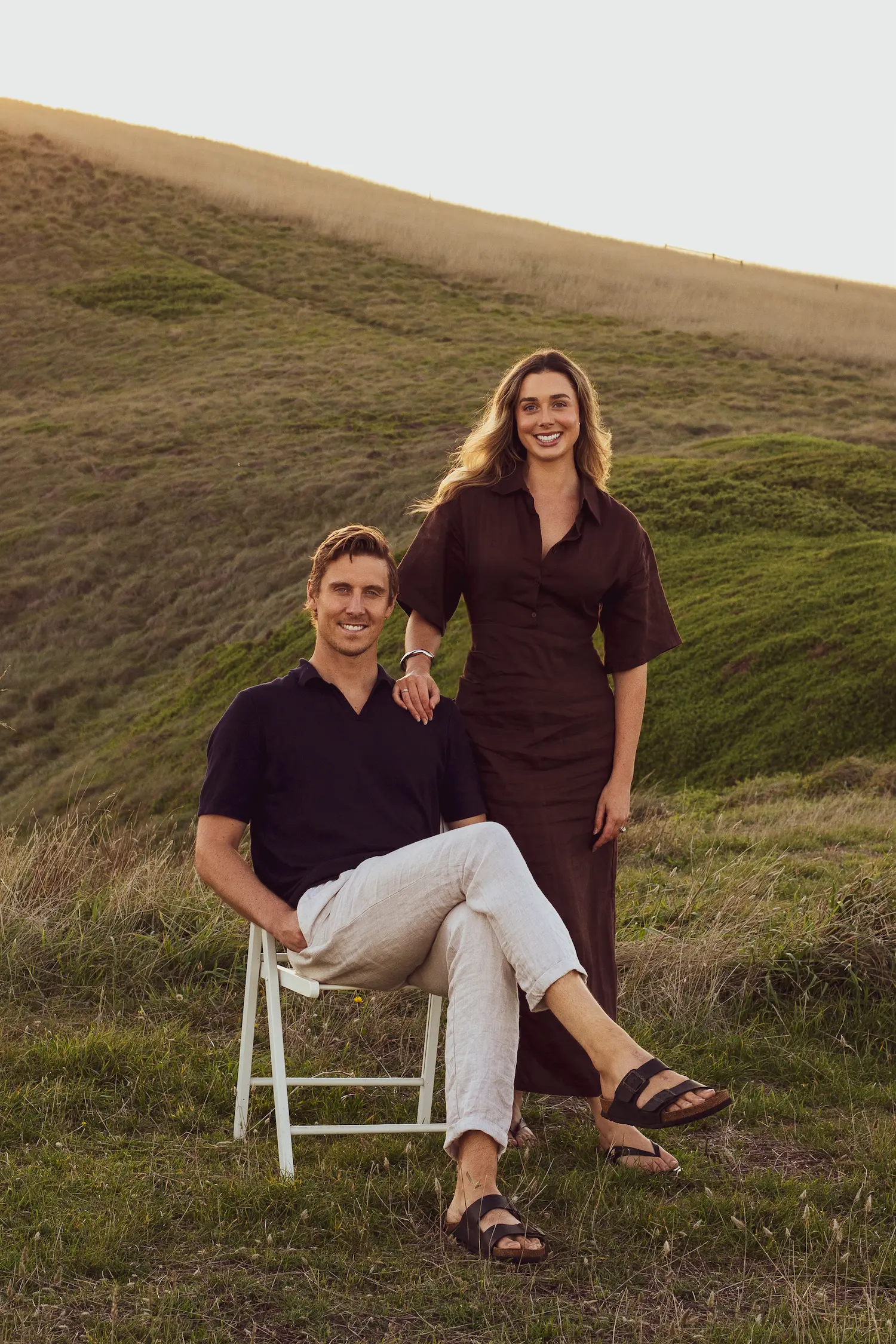 Smiling man sitting on white chair with crossed legs and woman standing beside in grassy hillside.