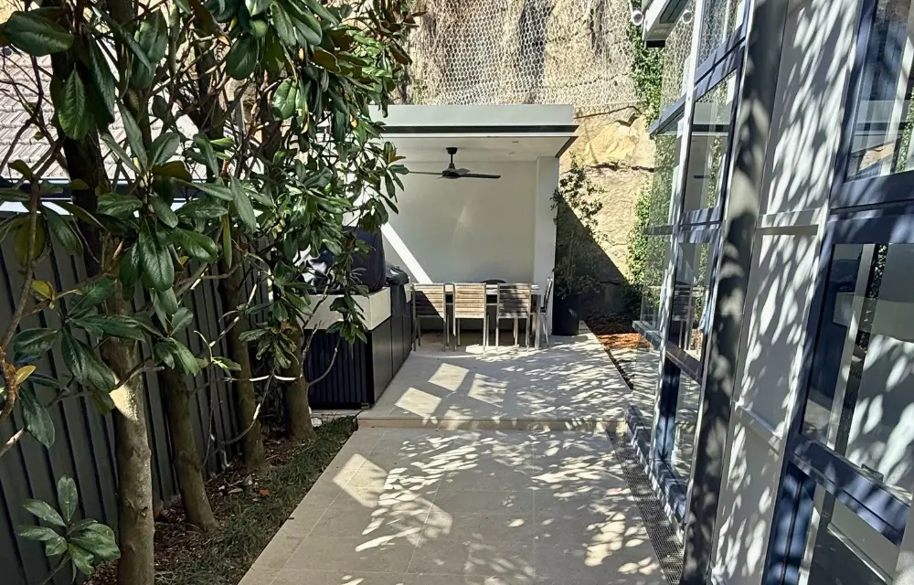 Outdoor patio with dining table and chairs under shade, adjacent to large windows and trees casting shadows.