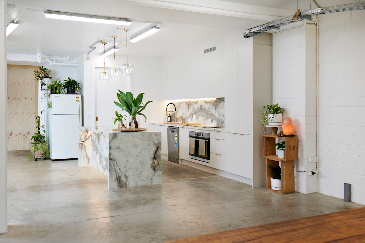 Modern kitchen with marble island, white cabinets, stainless oven, plants, and concrete floor.