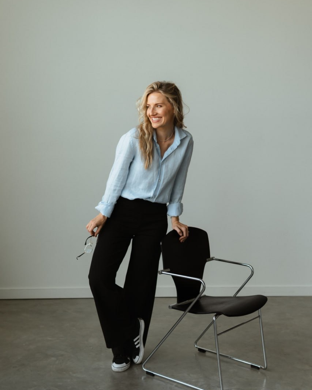 Smiling woman in light blue shirt and black pants leaning on a black chair with chrome legs.