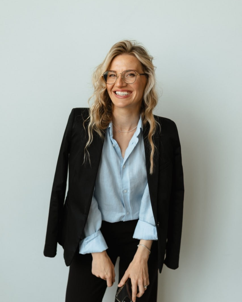 Smiling woman with blonde hair, glasses, light blue shirt, and black blazer against a plain background.