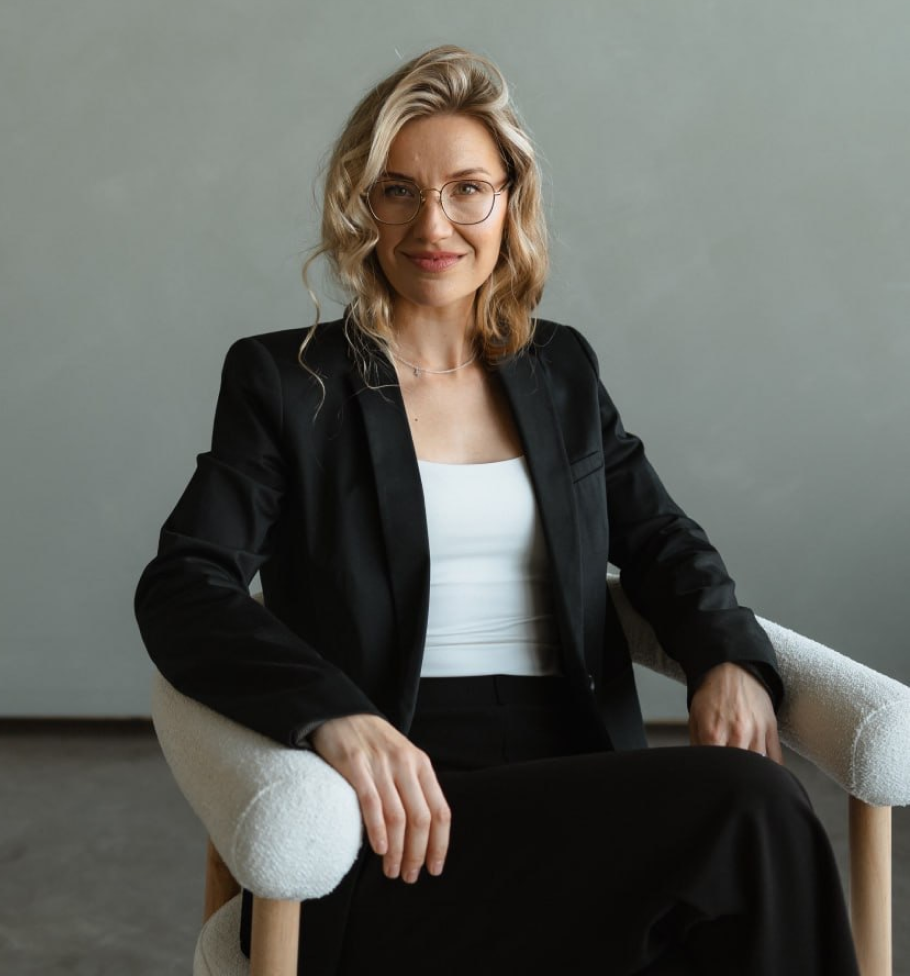 Smiling woman with blonde hair and glasses wearing black blazer and white top sitting on a white chair.