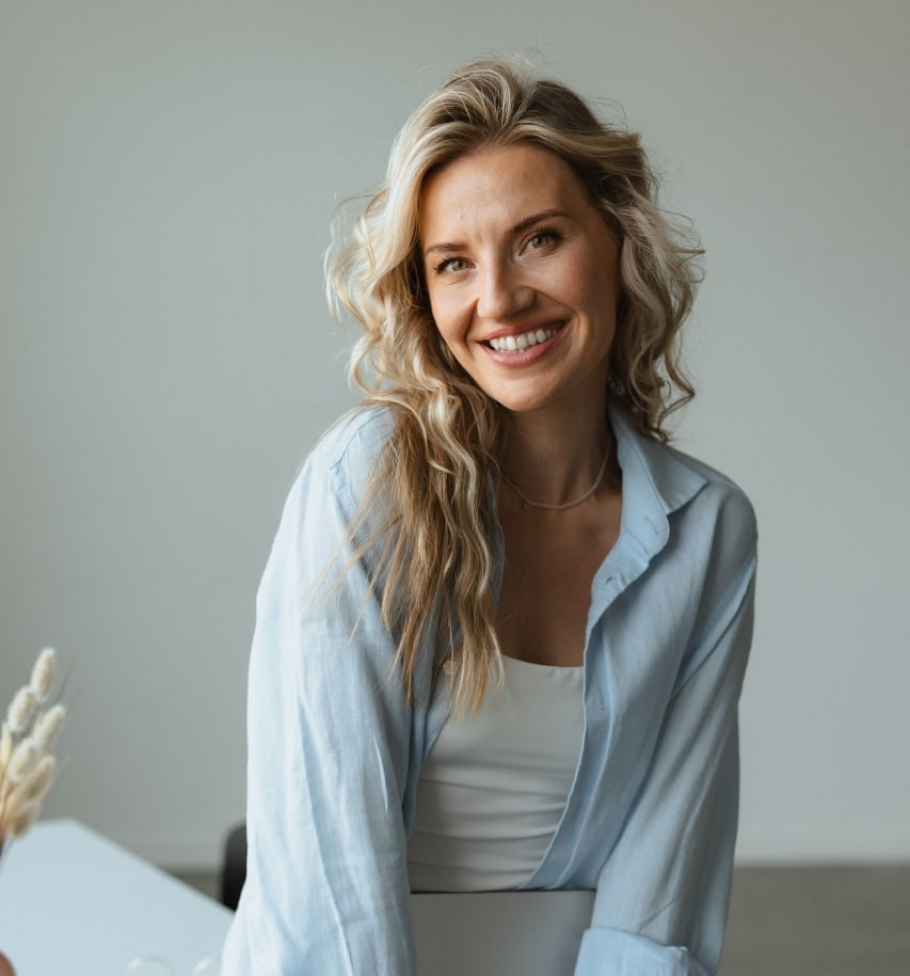 Smiling woman with long curly blonde hair wearing a light blue shirt over white top indoors.
