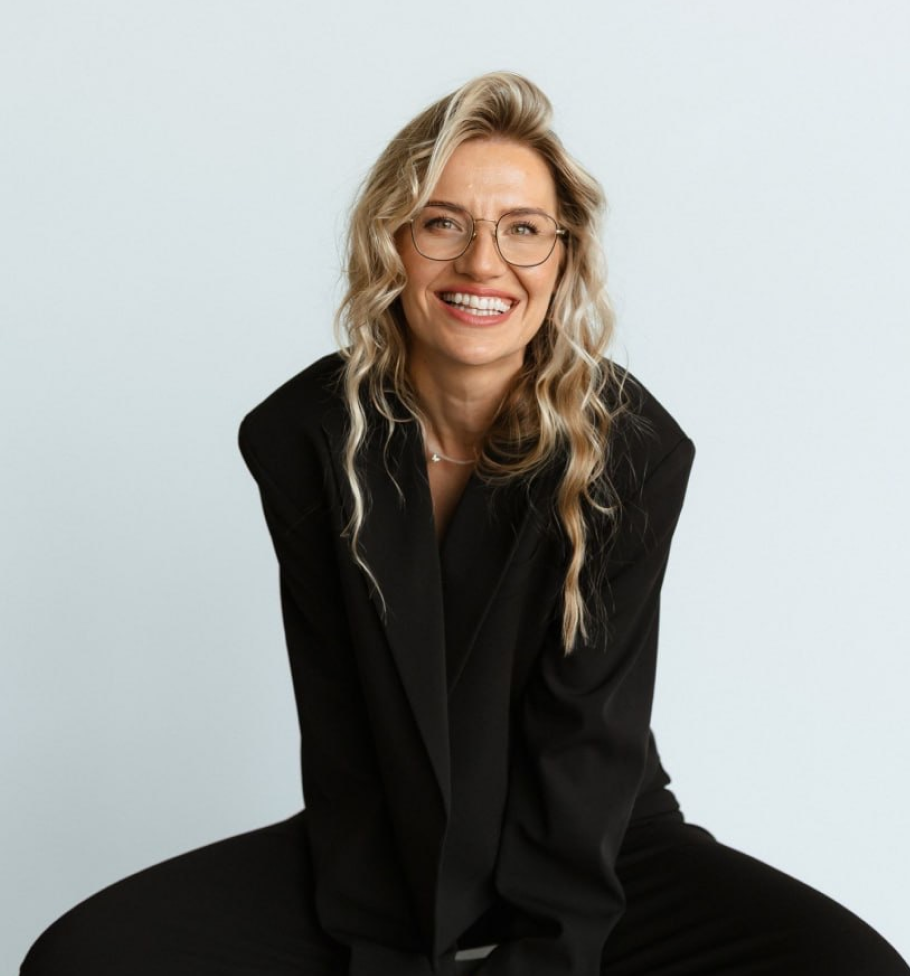 Smiling woman with wavy blonde hair and glasses wearing a black blazer against a plain background.