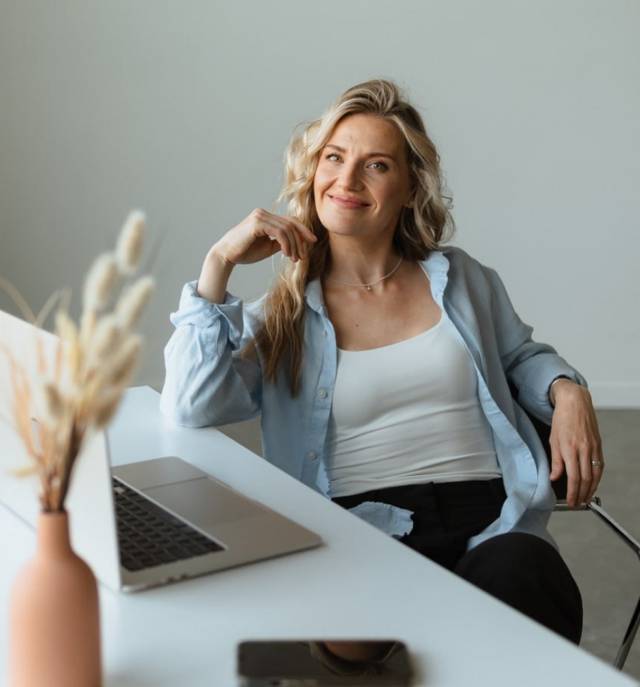 Smiling woman sitting at a desk with a laptop, phone, and vase with dried flowers.