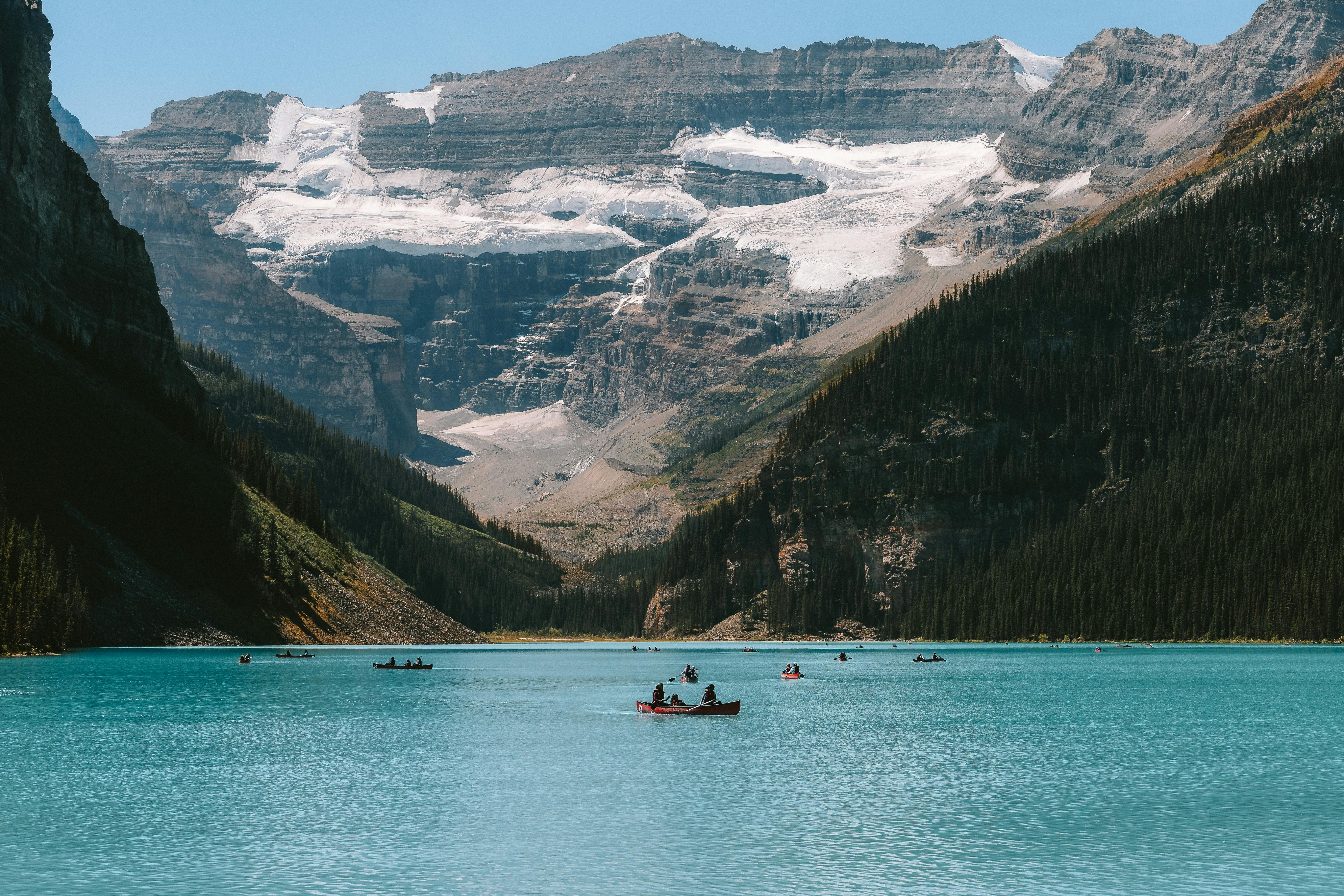 People canoeing on a turquoise lake surrounded by forested mountains and a glacier under a clear sky.