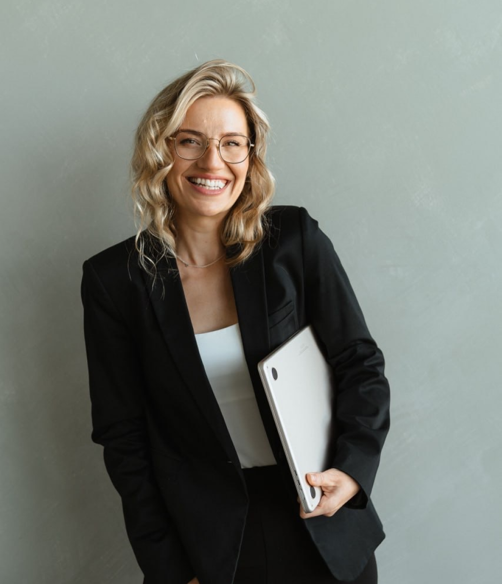 Smiling woman in glasses and black blazer holding a silver laptop against a gray wall.