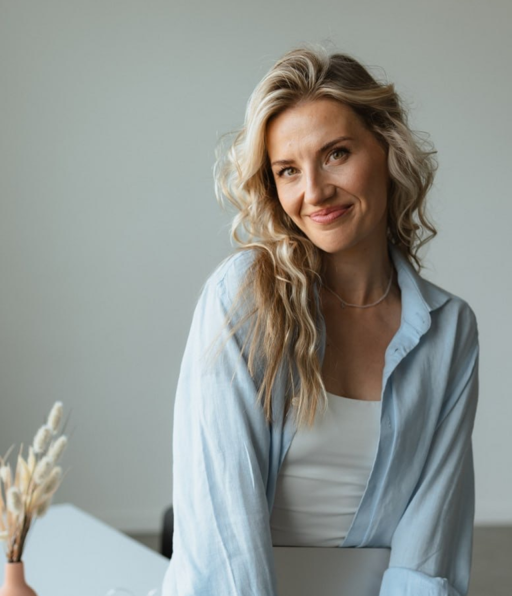 Smiling woman with long wavy blonde hair wearing a light blue shirt over a white top indoors.