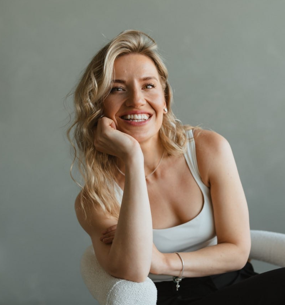 Smiling blonde woman in a white tank top sitting on a white chair against a grey background.