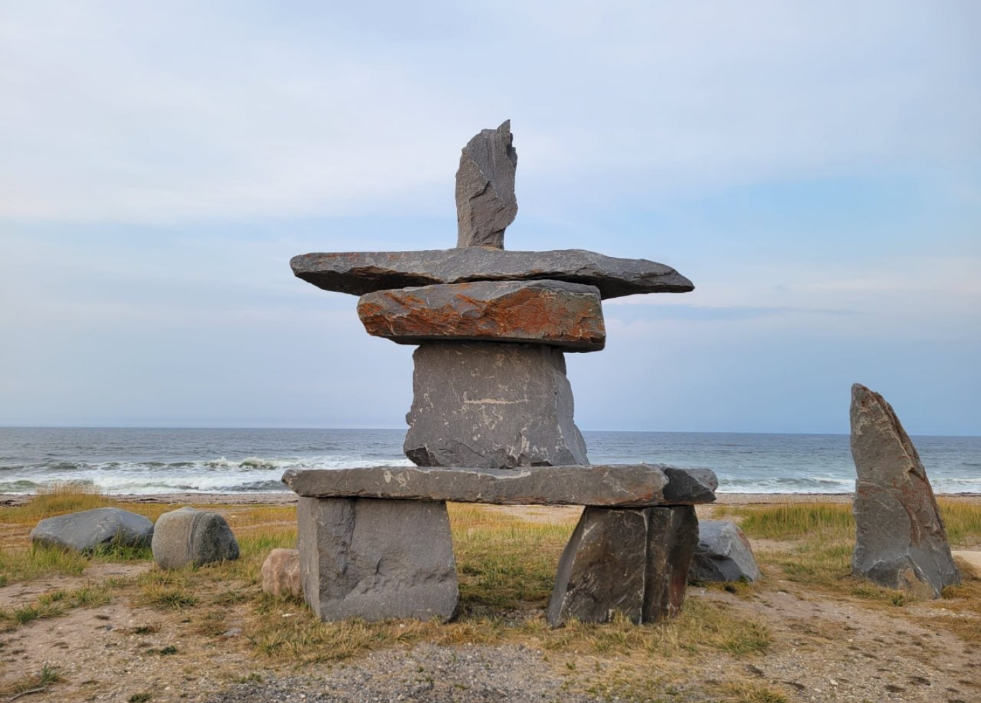 Stacked flat stones forming an inuksuk sculpture on grass near an ocean shore under a cloudy sky.