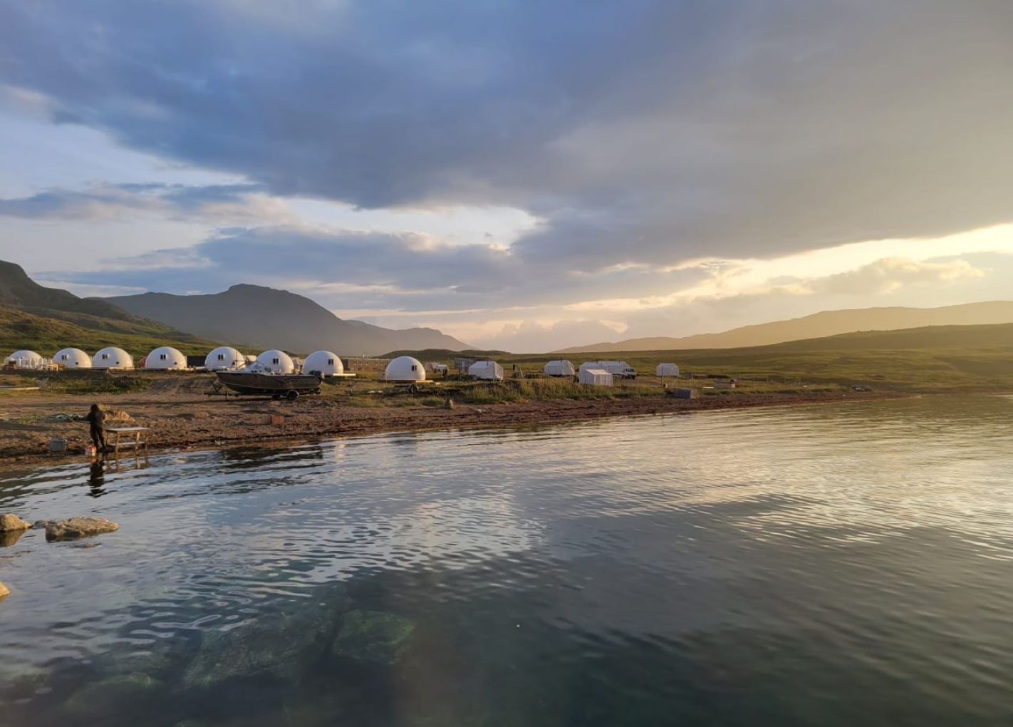 Waterfront view of a campsite with white dome tents, a boat, and mountains under a cloudy sky.