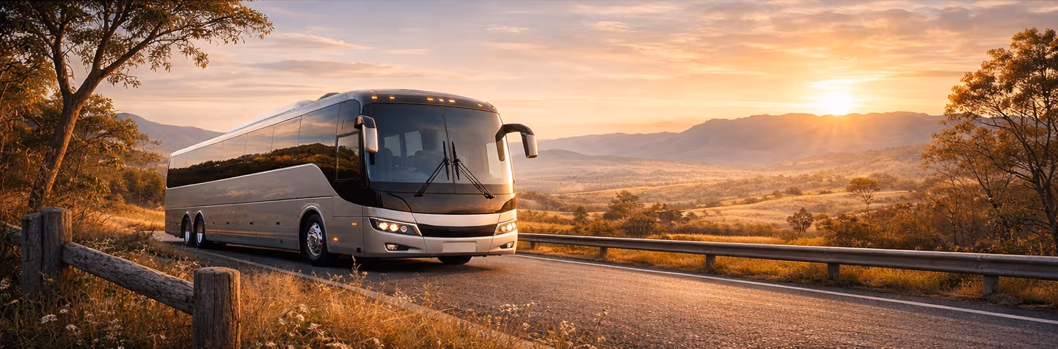 Modern coach bus driving on a rural road at sunset with mountains in the background.