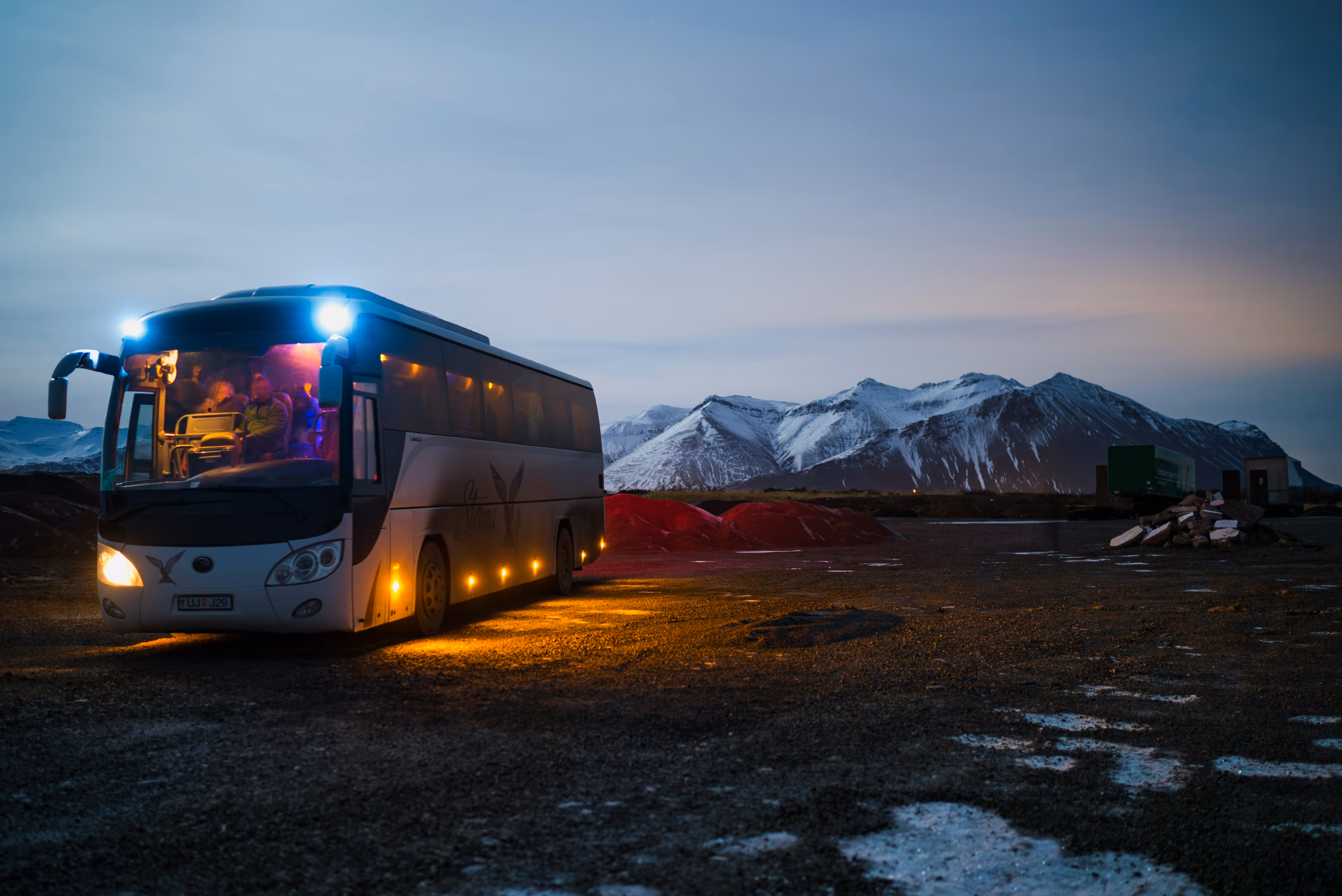 Tour bus with headlights on parked on a gravel area with snowy mountains in the background at dusk.