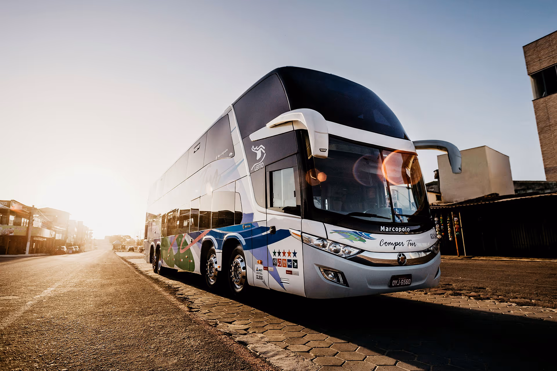 White and blue double-decker Marcopolo bus parked on a city street at sunset.