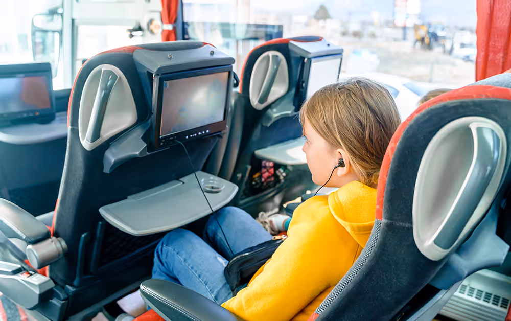 Child wearing a yellow hoodie and headphones sitting on a bus seat watching a screen on the seat in front.