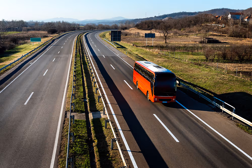 Red coach bus driving alone on a sunny multi-lane highway surrounded by grassy fields.