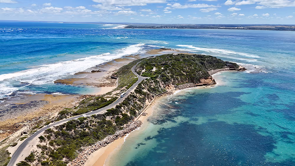 Aerial view of a coastal peninsula with a winding road surrounded by blue ocean and sandy shores under a partly cloudy sky.