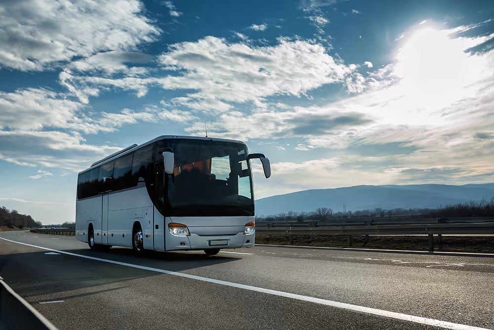White coach bus driving on a highway under a partly cloudy sky with hills in the background.