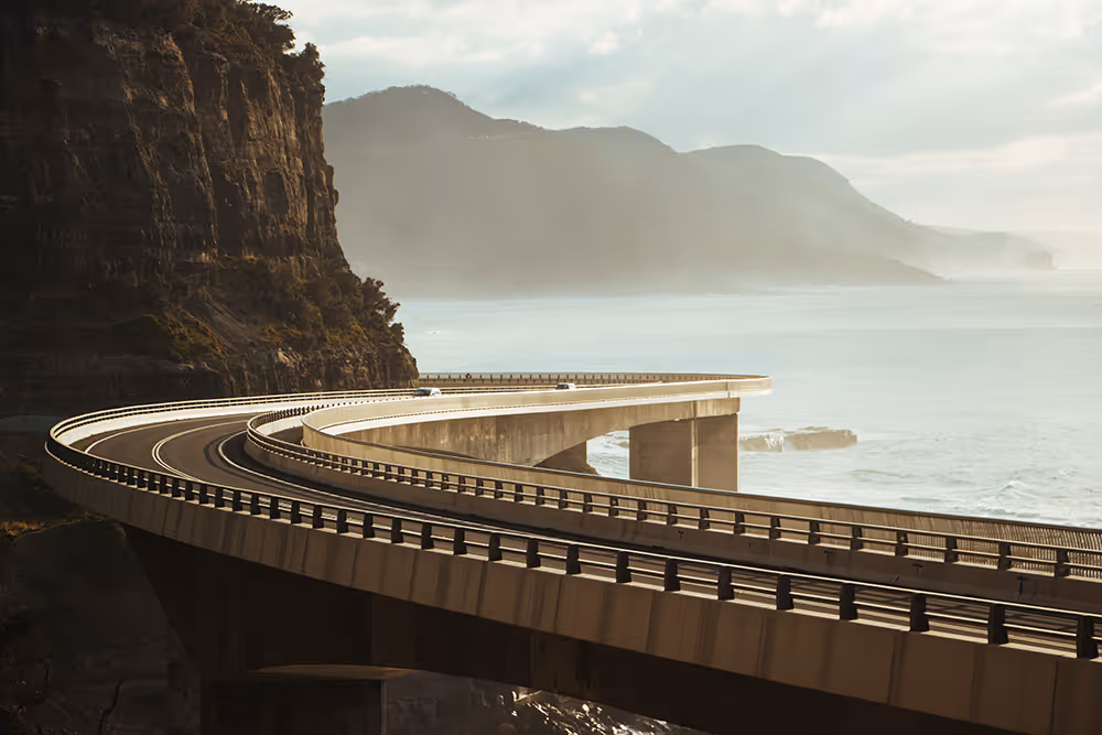 Curving coastal bridge with guardrails beside cliff over ocean under cloudy sky.
