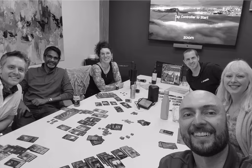 Six people smiling around a table playing the board game 7 Wonders with cards and coins, with a TV screen showing 'Tap Controller to Start' in the background.