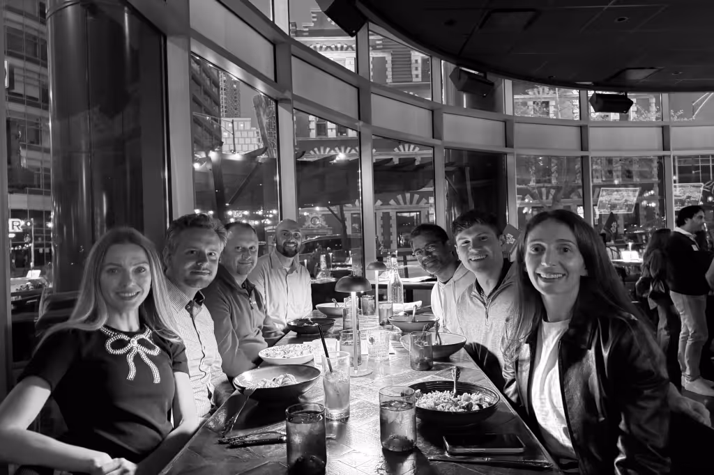 Seven people smiling and sitting around a table with food and drinks inside a restaurant with large windows.