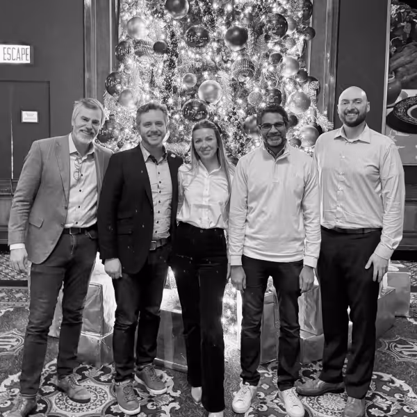 Group of five people standing and smiling in front of a decorated Christmas tree with wrapped gifts.