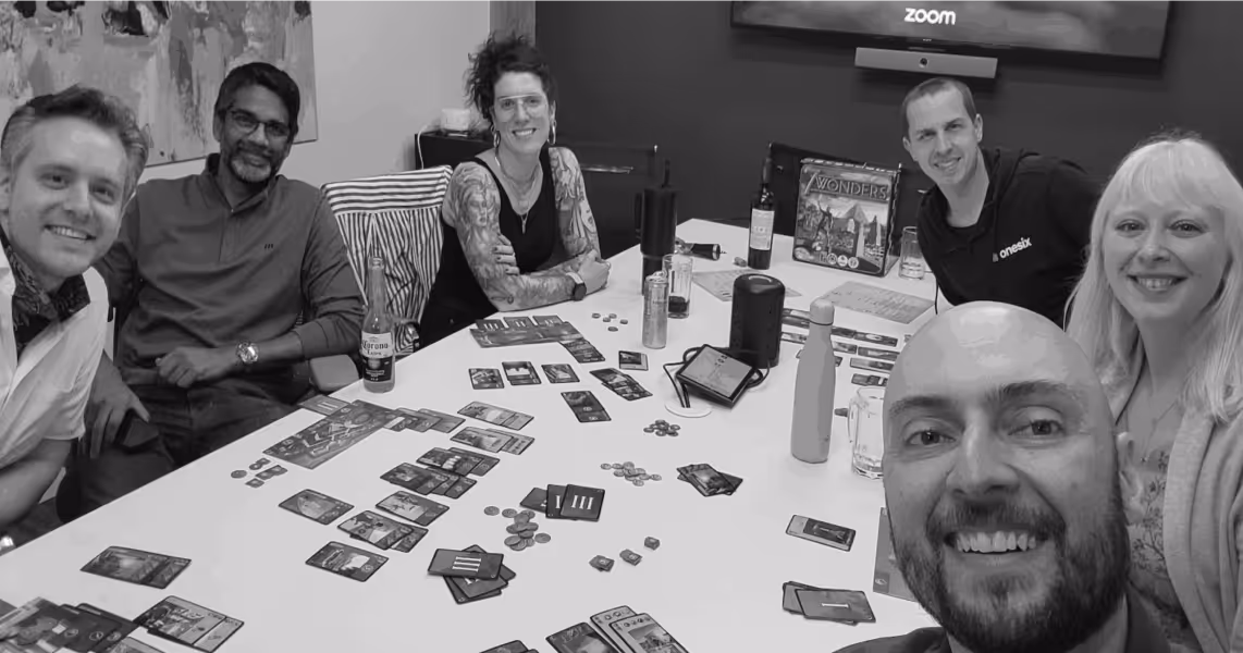 Group of six people smiling around a table playing the board game '7 Wonders' with cards and coins spread out.