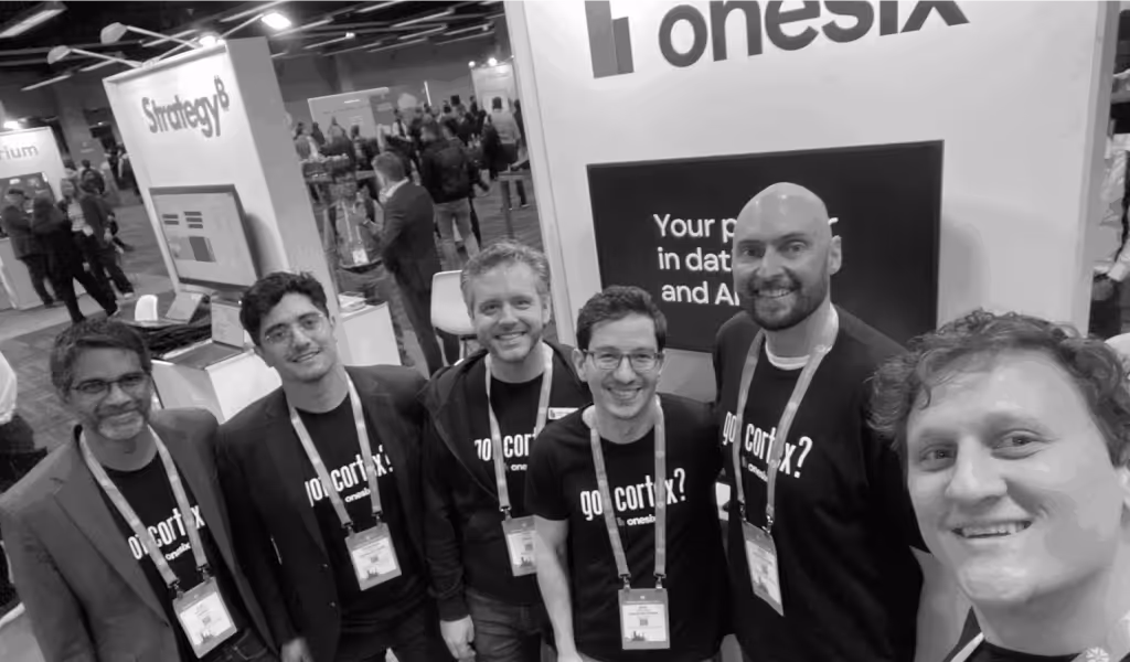 Six men wearing black onesix shirts and conference badges posing for a group selfie at a tech conference booth.