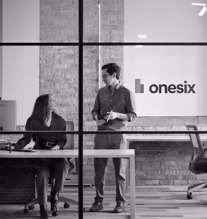 A man standing and holding a tablet talks to a seated woman at a desk in an office with a 'onesix' logo on the wall.