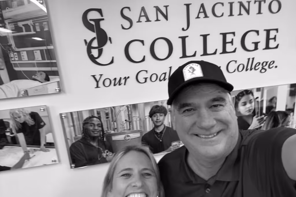 Two smiling adults taking a selfie in front of a San Jacinto College banner with student photos.