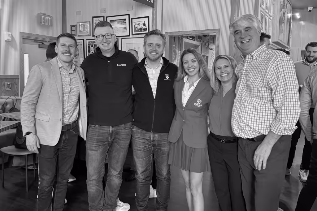 Group of six adults smiling and posing together inside a room with framed pictures on the wall.