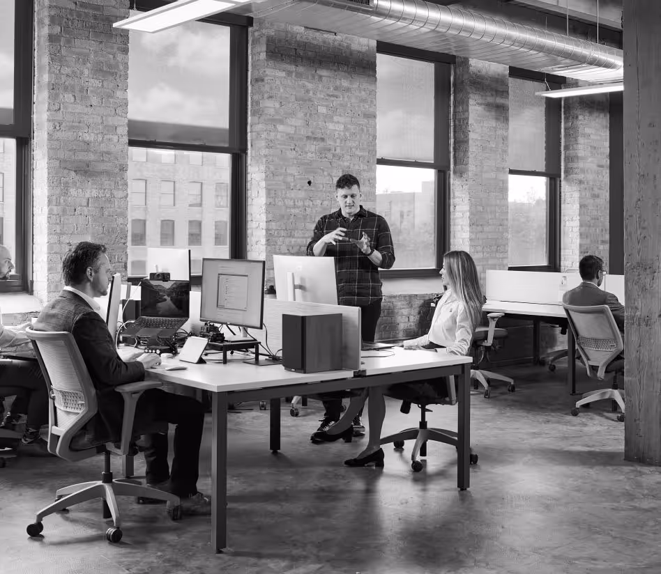 Four coworkers in a modern office, one standing and explaining while others sit at desks with computers.