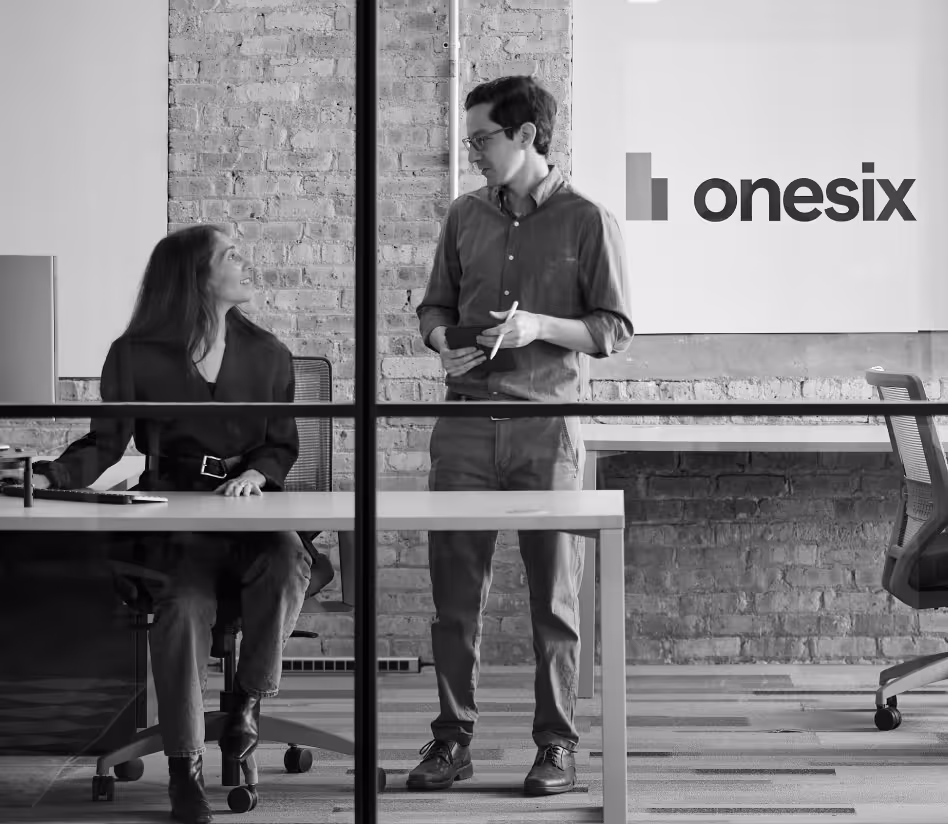 Woman sitting at desk and man standing, talking in modern office with exposed brick wall.