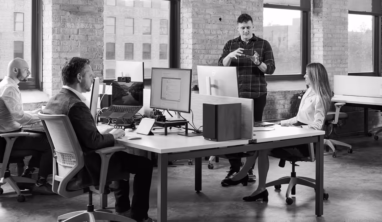 Four professionals in modern office with computers, one standing explaining something to seated colleagues.