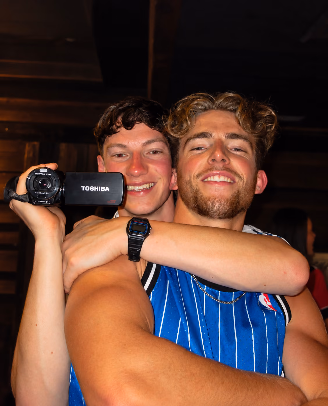 Two AmeriCamp summer camp friends smiling and hugging while holding a video camera at a camp social event.