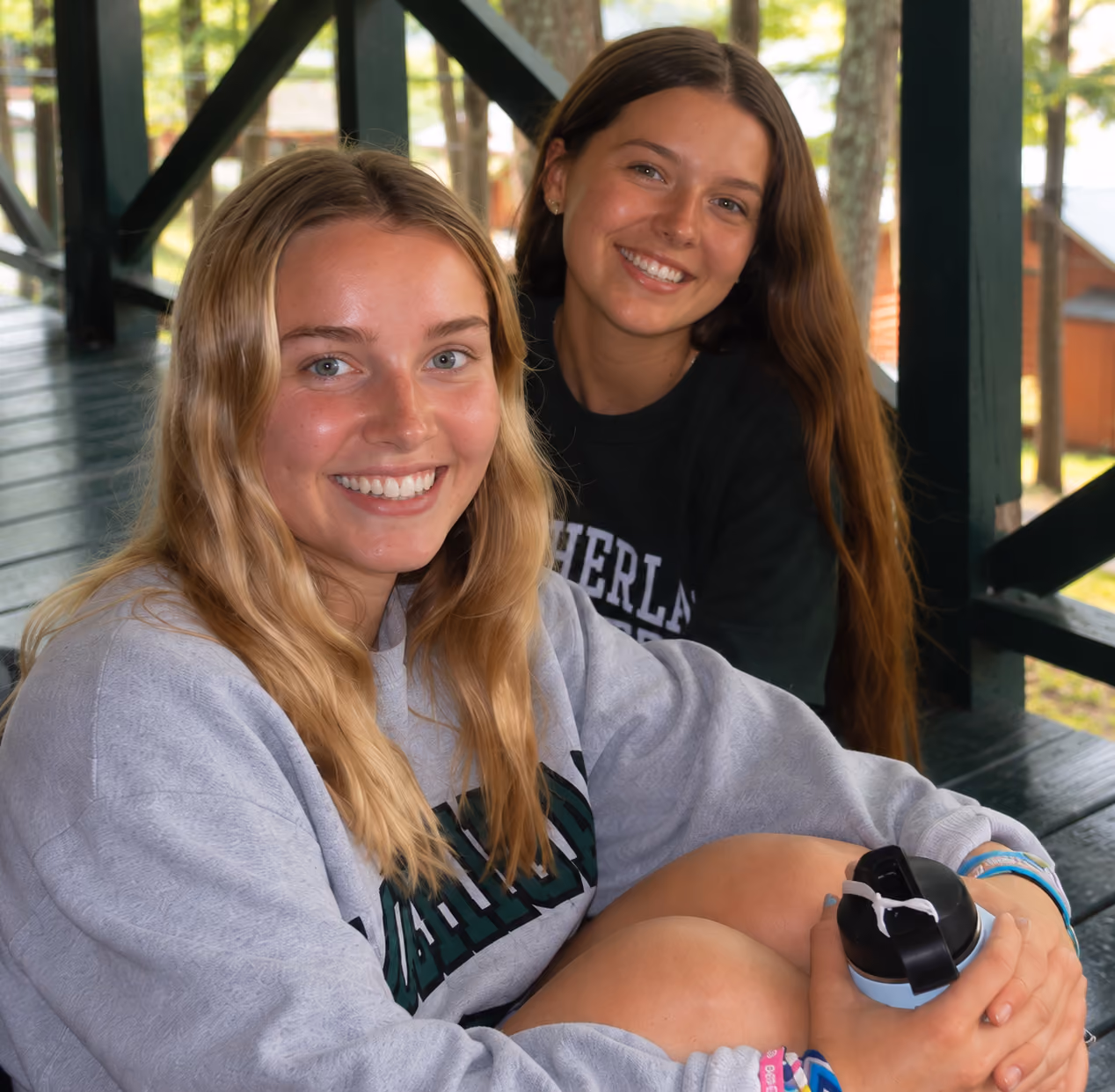 Two AmeriCamp summer camp friends smiling together on a wooden cabin porch in the USA.