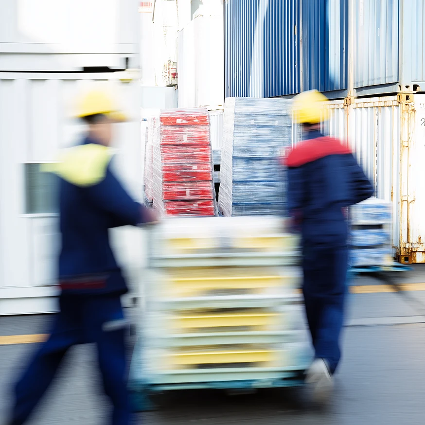 Two workers in safety helmets moving pallets stacked with goods in an industrial storage area with shipping containers.