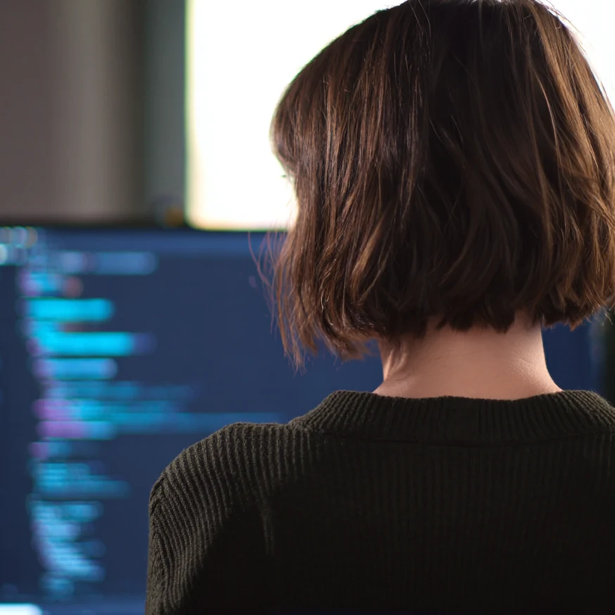 Person with short brown hair viewed from behind, working on a computer screen displaying blurred lines of code.
