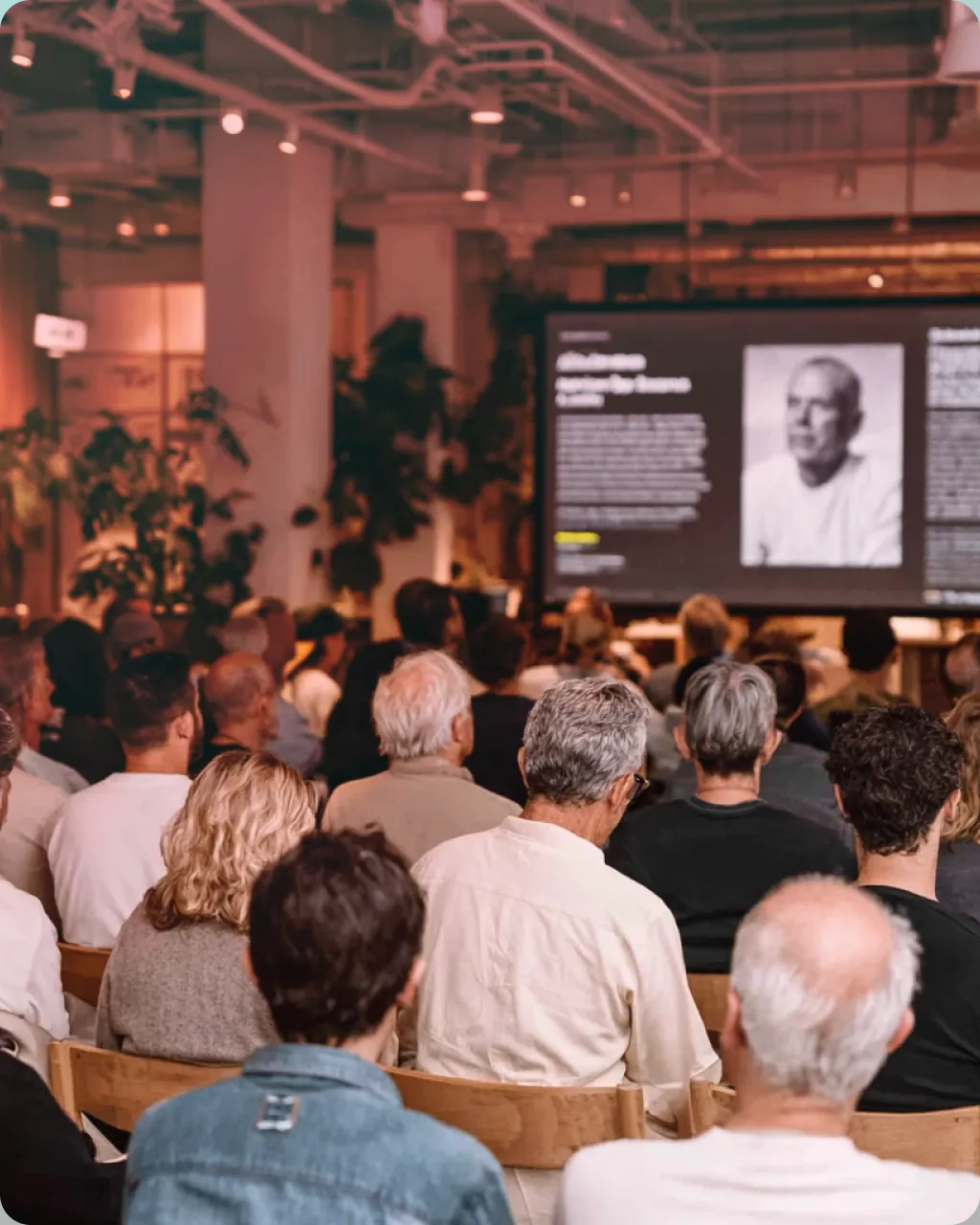 Audience seated in a conference room watching a presentation with a large screen displaying a black and white portrait and text.