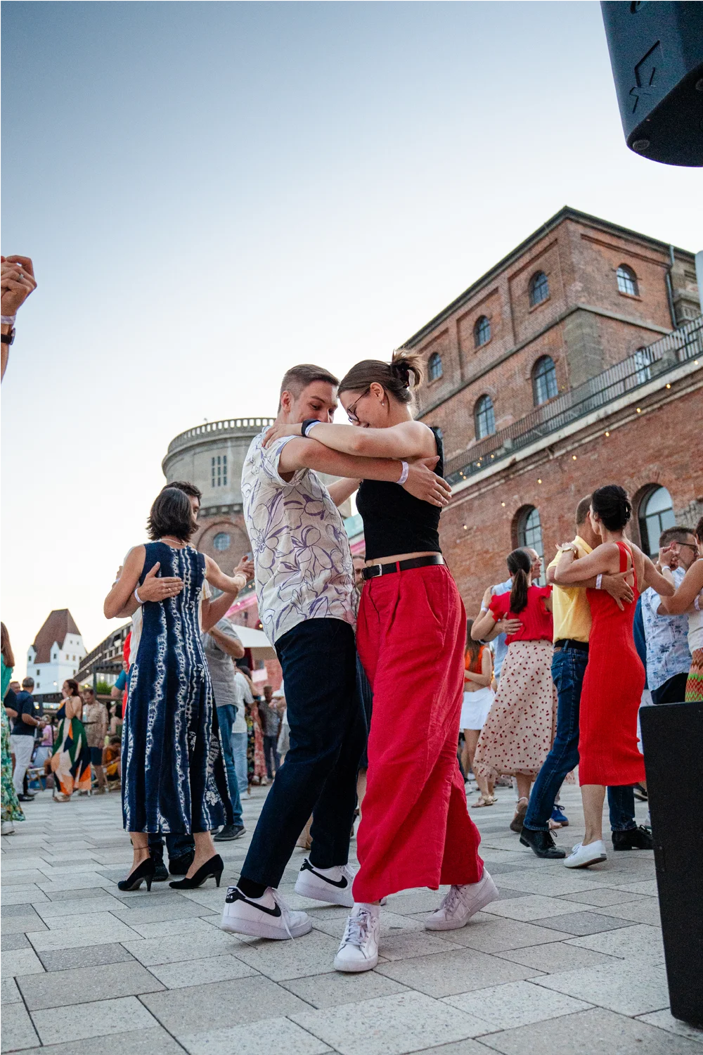 Paar tanzt eng zusammen bei einem Outdoor-Tanz-Event vor einem Backsteingebäude im Abendlicht.