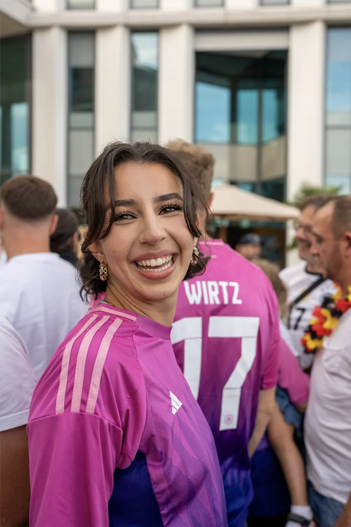 Junge Frau lächelt in einem pinken Adidas-Trikot auf einer Veranstaltung mit mehreren Menschen im Hintergrund.