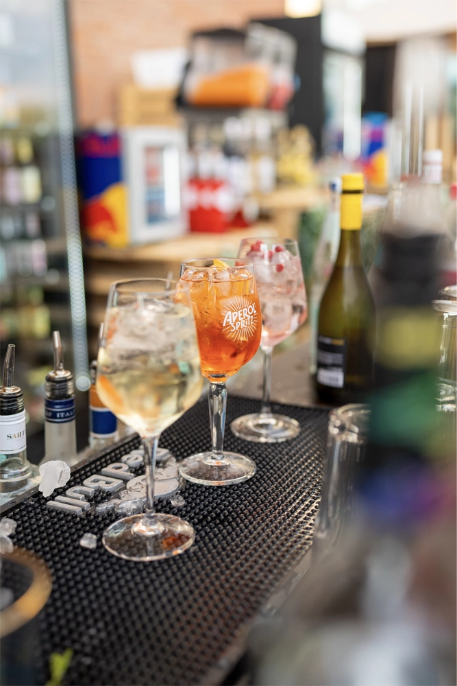 Drei Gläser auf einer Bar-Matte, in der Mitte ein Glas mit Aperol Spritz, umgeben von Eiswürfeln und einer Weinflasche im Hintergrund.