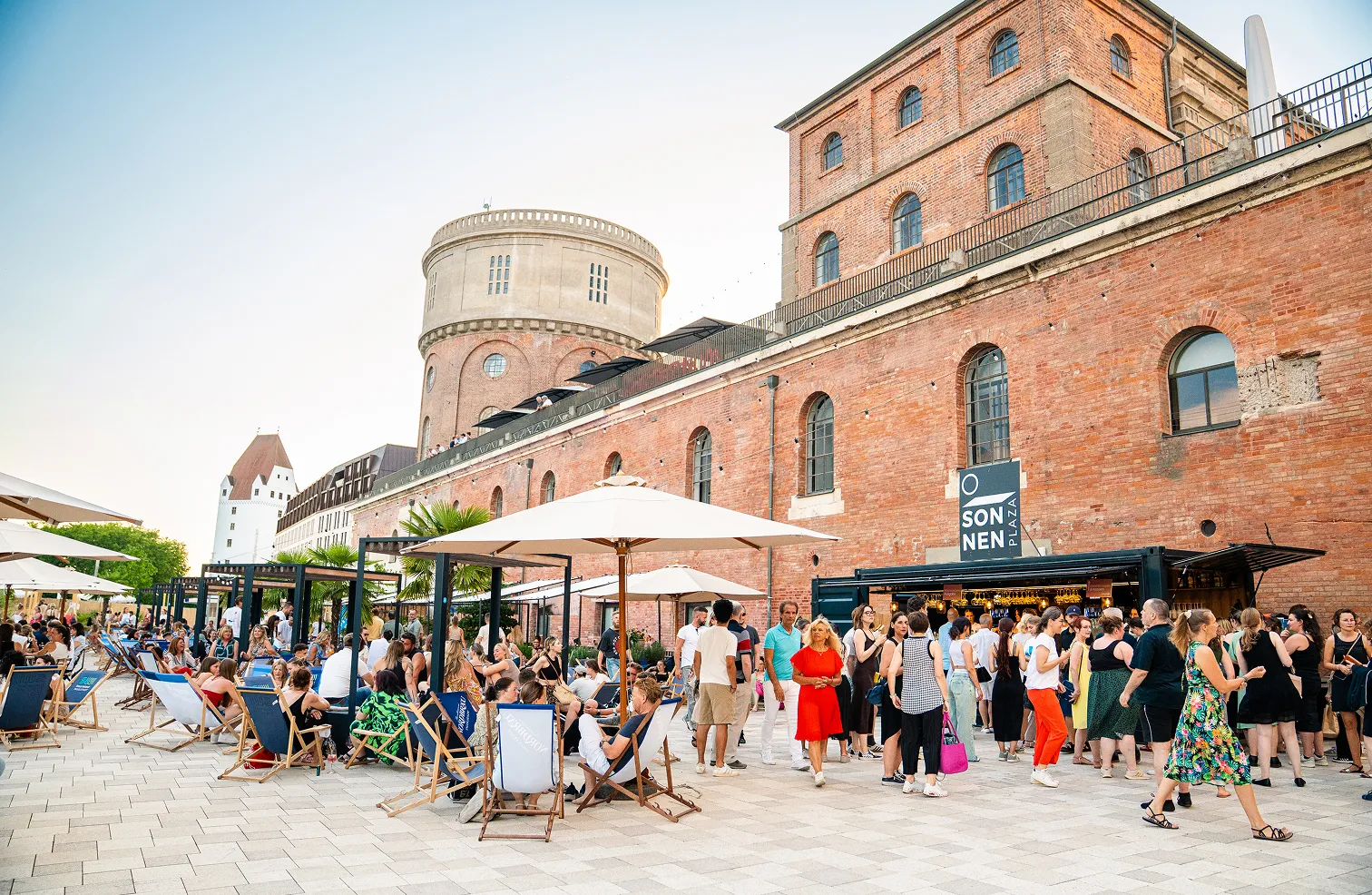 Menschen sitzen und stehen unter Sonnenschirmen vor einem historischen Backsteingebäude mit einem runden Turm bei klarem Himmel.