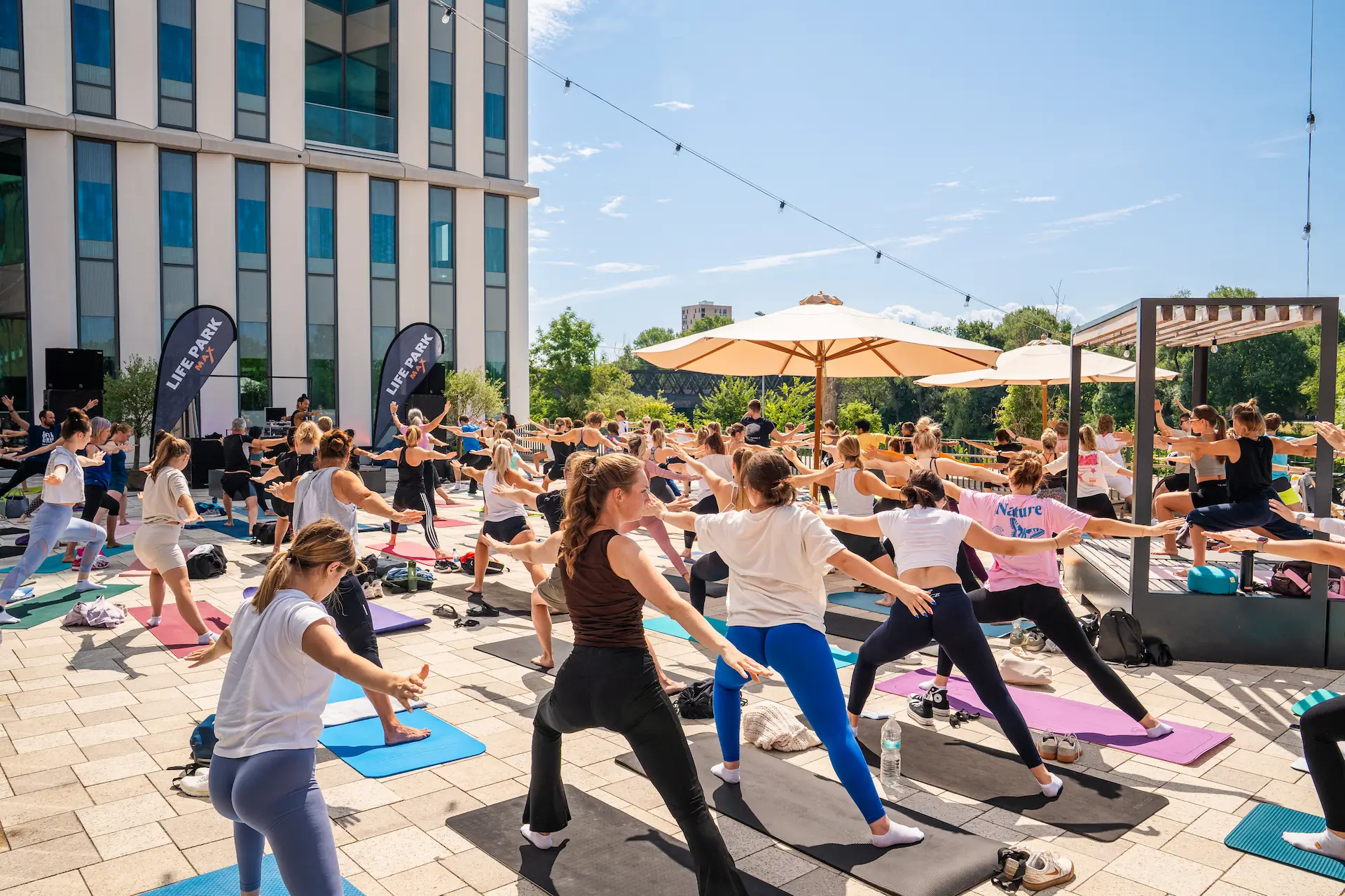 Eine große Gruppe von Menschen macht Yoga im Freien auf Yogamatten vor einem modernen Gebäude an einem sonnigen Tag.