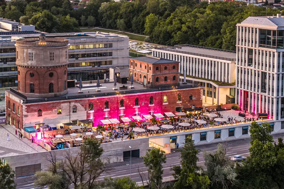 Belebter urbaner Platz mit beleuchteten Gebäuden, Außenbereich mit Sitzgelegenheiten und Menschen, umgeben von Straßen und Bäumen.