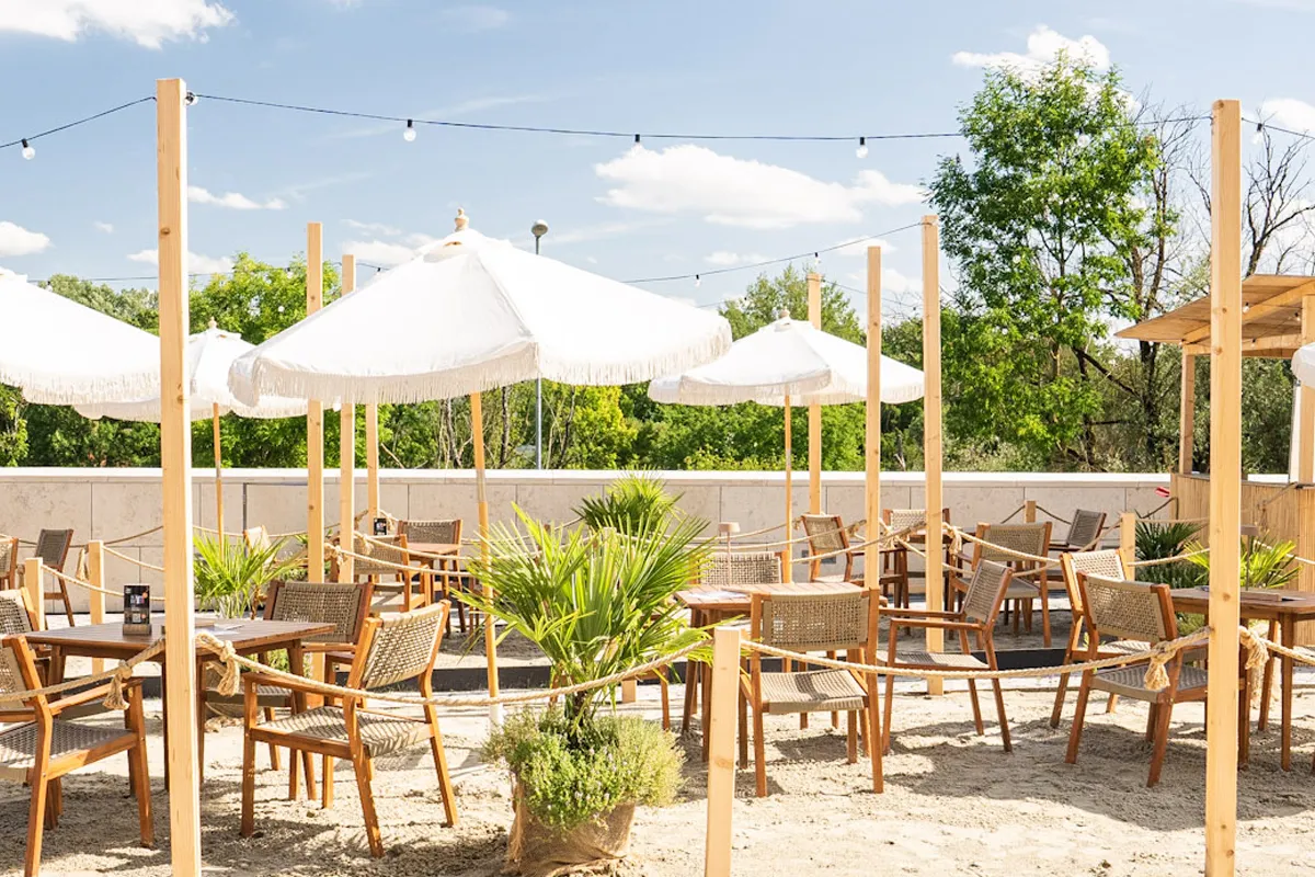 Außenbereich eines Cafés oder Restaurants mit Stühlen und Tischen auf Sand, weißen Sonnenschirmen und grünen Pflanzen unter blauem Himmel.