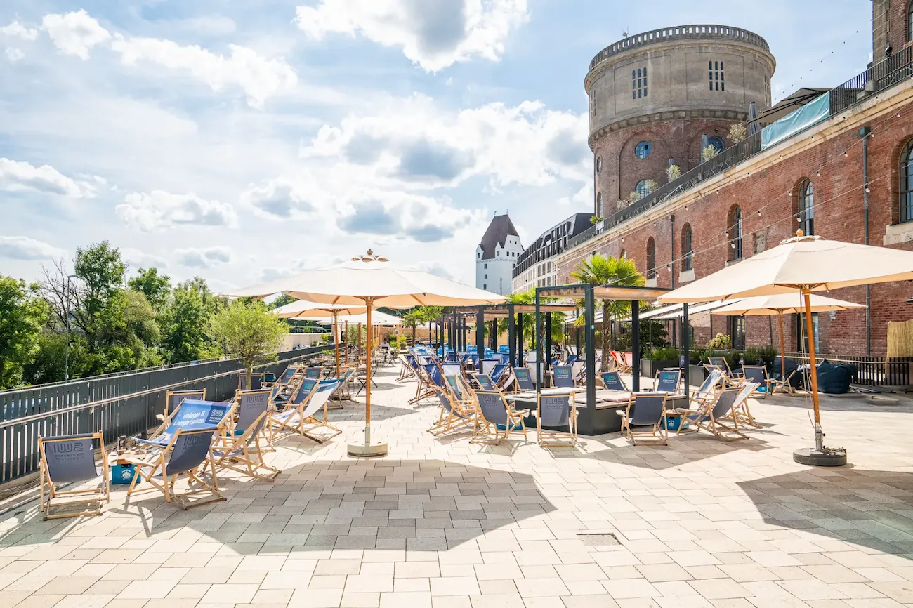 Große Terrasse mit Liegestühlen und Sonnenschirmen vor einem historischen Backsteingebäude unter blauem Himmel.